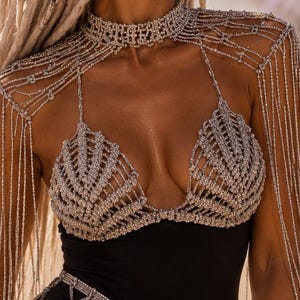 May include: A black bodysuit adorned with an intricate silver beaded harness that features a high choker neckline, shell-shaped bust detailing, and long, cascading fringe sleeves. A matching beaded net skirt overlay drapes over the hips.