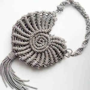 May include: Silver-colored, handmade handbag with a unique spiral design. The bag features a tassel and a braided strap. The bag is made of a metallic material and has a heart-shaped clasp. The bag is on a white background.