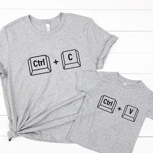 May include: Two gray t-shirts, one adult and one child size, with black keyboard key graphics. The adult shirt has "Ctrl + C" and the child's shirt has "Ctrl + V". Both shirts are on a white wooden surface.