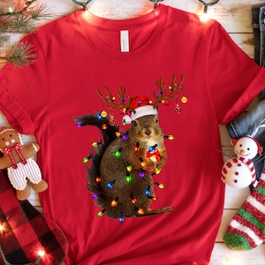 May include: A red t-shirt featuring a squirrel wearing a Santa hat and antlers, wrapped in Christmas lights. The image is set against a festive backdrop with a Christmas stocking, gift, jeans, and socks.