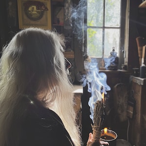 K&ouml;nnte beinhalten: Eine Person mit langen wei&szlig;en Haaren h&auml;lt ein brennendes Salbeib&uuml;ndel, das blaue Rauchschwaden erzeugt. In der anderen Hand wird eine kleine Tasse mit einer Flamme gehalten. Die Szene spielt in einem schwach beleuchteten Raum mit einem Fenster im Hintergrund.