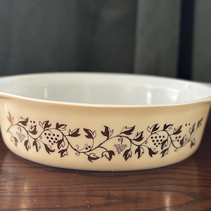 Puede incluir: Un plato para hornear ovalado blanco con un diseño de vid y uvas marrones. El plato tiene un interior blanco liso y un borde ligeramente elevado.