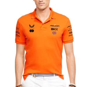 Może przedstawiać: Pomarańczowa koszulka polo z kołnierzykiem i krótkimi rękawami. Koszulka posiada wiele logo, w tym "McLaren" i "OKX", oraz małą flagę Union Jack. Koszulka jest połączona z białymi spodniami i paskiem w paski.