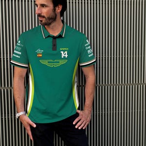 Könnte beinhalten: Grünes Poloshirt mit einem weißen und gelben Design. Das Shirt trägt die Nummer 14 und das Wort "Aramco" auf der Vorderseite. Das Shirt hat auch die spanische Flagge und ein Logo mit Flügeln.