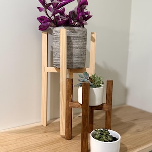 Puede incluir: Dos soportes de plantas de madera, uno natural y otro teñido de marrón, cada uno con una planta en maceta. El soporte más alto sostiene una maceta gris texturizada con una planta morada y verde. El soporte más pequeño sostiene una maceta blanca con una planta verde.