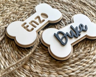 May include: Two white wooden dog bone-shaped tags with black lettering. The tag on the left says "Enzo" and the tag on the right says "Duke".