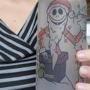 May include: Frosted glass can-shaped tumbler with a bamboo lid. The tumbler features a festive design of Jack Skellington in a Santa Claus outfit, holding Christmas lights. A small green creature and the text "Ms. Alvarado" are also visible.