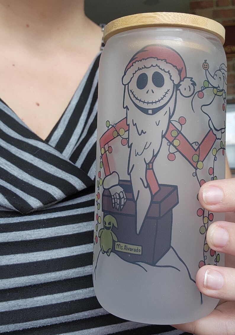 May include: Frosted glass can-shaped tumbler with a bamboo lid. The tumbler features a festive design of Jack Skellington in a Santa Claus outfit, holding Christmas lights. A small green creature and the text "Ms. Alvarado" are also visible.