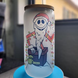 May include: A frosted glass can-shaped tumbler with a wooden lid. The tumbler features a cartoon illustration of a character dressed as Santa Claus, with a long white beard and a red Santa hat. The design includes a string of Christmas lights and the text "Ms Alvarado".