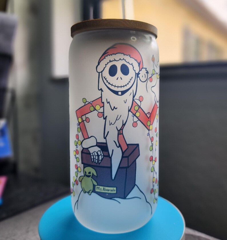 May include: A frosted glass can-shaped tumbler with a wooden lid. The tumbler features a cartoon illustration of a character dressed as Santa Claus, with a long white beard and a red Santa hat. The design includes a string of Christmas lights and the text "Ms Alvarado".