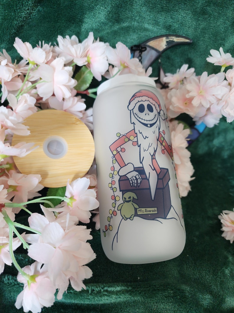 May include: A frosted glass tumbler with a bamboo lid. The tumbler features a cartoon illustration of Jack Skellington in a Santa hat, with a beard and a gift box. The design includes a small Oogie Boogie character and the text "Ms. Alvarado".