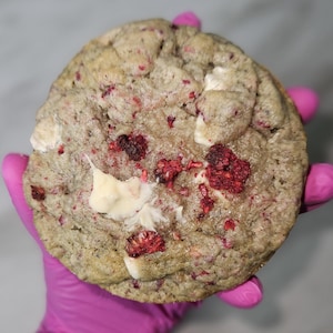 May include: A large, round cookie with a light brown base, studded with bright red raspberries and white chocolate chunks. The cookie is held by a person wearing a pink glove. The cookie appears freshly baked and has a slightly uneven texture.