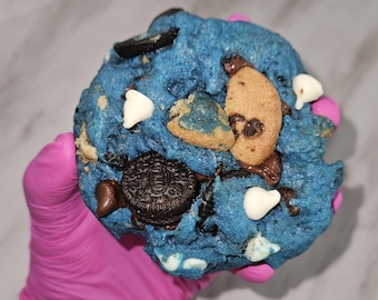 May include: A large, blue-colored cookie with various toppings. The cookie features Oreo pieces, white chocolate chips, and other cookie pieces. The cookie is held by a person wearing a pink glove.