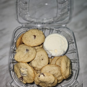 May include: A clear plastic container filled with small, round, brown chocolate chip cookies. A small, clear plastic container with white frosting is in the center of the container.