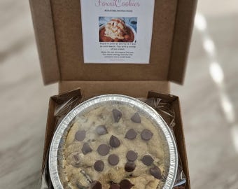 May include: A chocolate chip cookie in a silver foil pie pan, inside a brown cardboard pizza box. The cookie is topped with chocolate chips and is ready to be eaten.