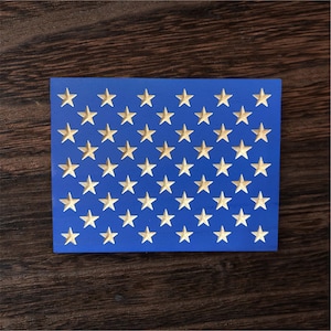 May include: A blue wooden block with 50 gold-colored stars carved into its surface. The stars are arranged in a grid pattern. The block is set against a dark brown wooden background.