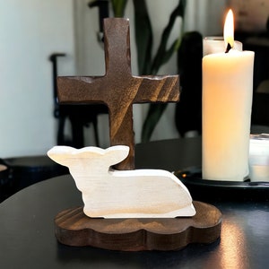 May include: A wooden cross with a white wooden lamb lying at the base. The cross is brown and the lamb is white. The cross and lamb are on a brown wooden base. The base has a scalloped edge. The cross and lamb are on a black table. There is a lit candle in the background.
