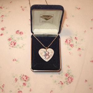 May include: A heart-shaped pendant necklace with the letter "B" in a floral design, displayed in a blue velvet jewelry box. The pendant is white with pink and blue floral accents. The box has a gold trim and a gold chain.