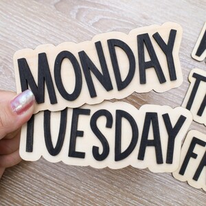 May include: Wooden day-of-the-week signs with black lettering. The days of the week are displayed on individual wooden cutouts. The word "MONDAY" and "TUESDAY" are in the foreground, with other days partially visible.
