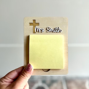 May include: A wooden memo holder with a cross cutout and the name "Mrs Smith" in white script. A yellow sticky note pad is attached to the holder. The holder is being held by a hand.