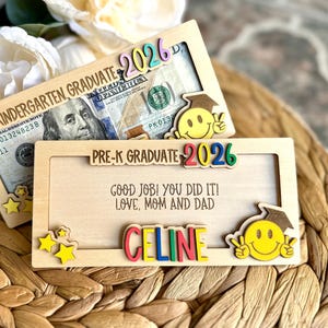May include: Two wooden graduation gift holders with a $100 bill inside. One says "Kindergarten Graduate 2026" and the other says "Pre-K Graduate 2026". Both have smiley faces and the name "CELINE".
