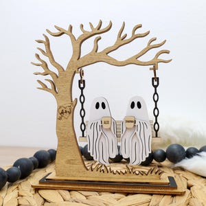 May include: Wooden Halloween decoration featuring a tree with a swing. Two white ghost figures sit on the swing, holding mugs. The tree has a heart with initials carved into it. The base is a wooden platform.