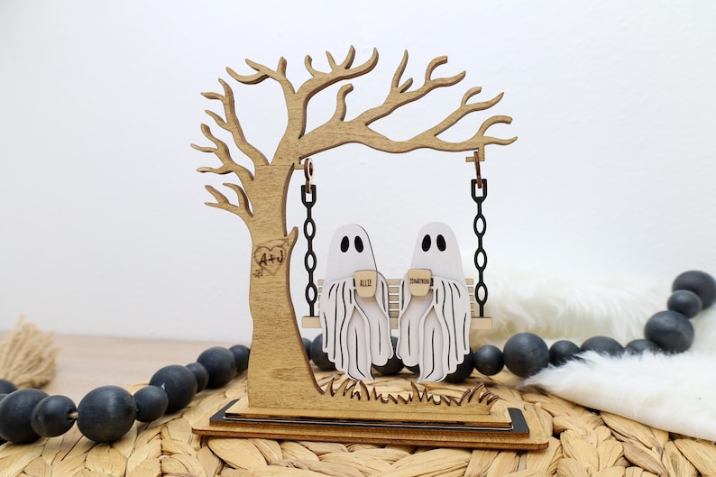 May include: Wooden Halloween decoration featuring a tree with a swing. Two white ghost figures sit on the swing, holding mugs. The tree has a heart with initials carved into it. The base is a wooden platform.