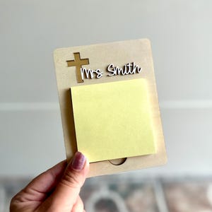 May include: A personalized notepad holder with a cross and the name "Mrs Smith" cut out of the top. The holder is made of light brown wood and has a yellow sticky note pad attached.