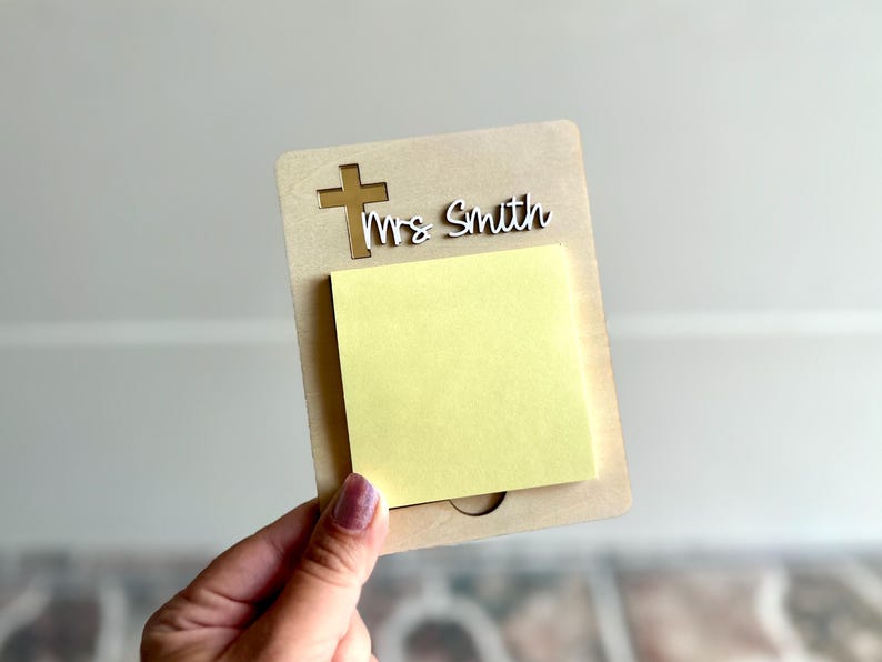 May include: A personalized notepad holder with a cross and the name "Mrs Smith" cut out of the top. The holder is made of light brown wood and has a yellow sticky note pad attached.