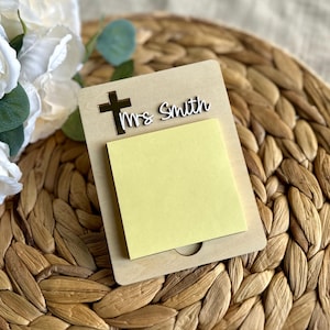 May include: A wooden memo pad with a cutout cross and the name "Mrs Smith" in white script. A yellow sticky note pad is attached. The memo pad is on a woven surface, with white flowers in the background.