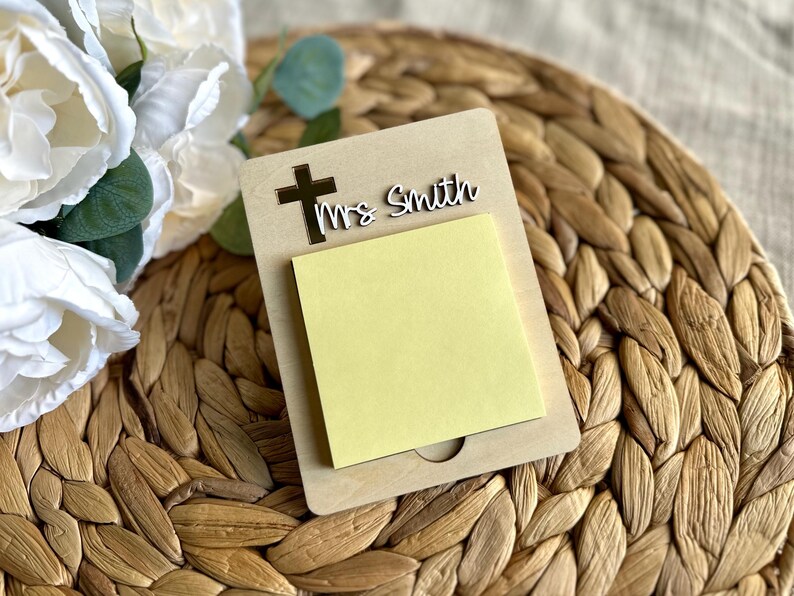 May include: A wooden memo pad with a cutout cross and the name "Mrs Smith" in white script. A yellow sticky note pad is attached. The memo pad is on a woven surface, with white flowers in the background.