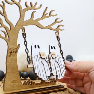 May include: A wooden Halloween decoration featuring a tree with a swing. Two white ghost figures are on the swing. The tree has a heart with the initials "A+J" carved into it. The base is a dark brown color.