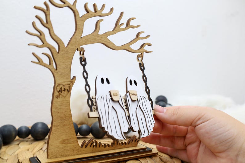 May include: A wooden Halloween decoration featuring a tree with a swing. Two white ghost figures are on the swing. The tree has a heart with the initials "A+J" carved into it. The base is a dark brown color.