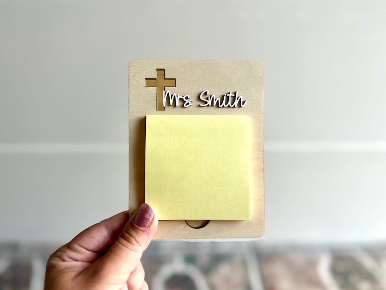 May include: A wooden memo holder with a cross cutout and the name "Mrs Smith" in white script. A yellow sticky note pad is attached to the holder. The holder is being held by a hand.
