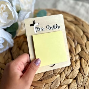 May include: A wooden memo pad with a cross cutout and the name "Ms. Smith" in script. A yellow sticky note pad is attached. The memo pad is held in front of a woven background and white flowers.