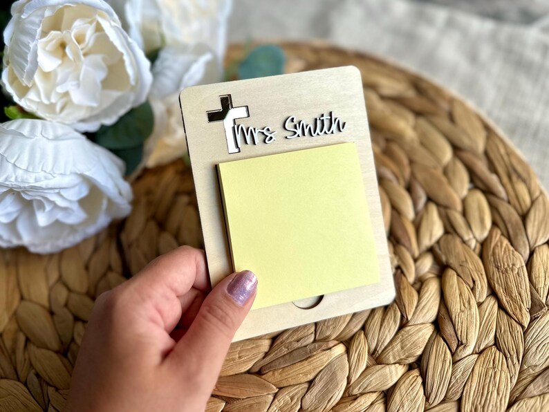 May include: A wooden memo pad with a cross cutout and the name "Ms. Smith" in script. A yellow sticky note pad is attached. The memo pad is held in front of a woven background and white flowers.