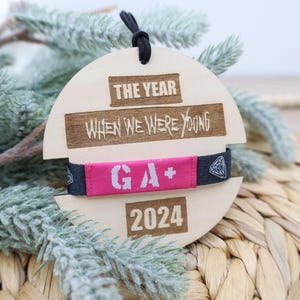 May include: A wooden ornament with the text "The Year When We Were Young" and a pink and black wristband with the text "GA+" and the year "2024".
