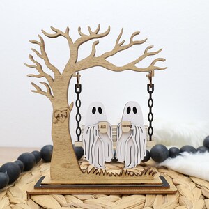 May include: Wooden Halloween decoration featuring a tree with a swing. Two ghost figures sit on the swing, holding mugs. The tree has a heart with initials carved into it. The base is rectangular and the overall color scheme is light brown and white.