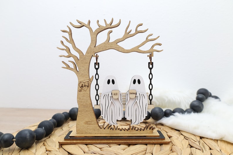 May include: Wooden Halloween decoration featuring a tree with a swing. Two ghost figures sit on the swing, holding mugs. The tree has a heart with initials carved into it. The base is rectangular and the overall color scheme is light brown and white.