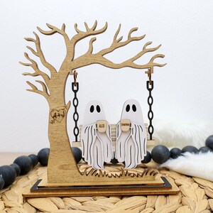 May include: Wooden Halloween decoration featuring a tree with a swing. Two ghost figures are seated on the swing, holding mugs. The tree has a heart with initials carved into it. The base is on a woven surface.