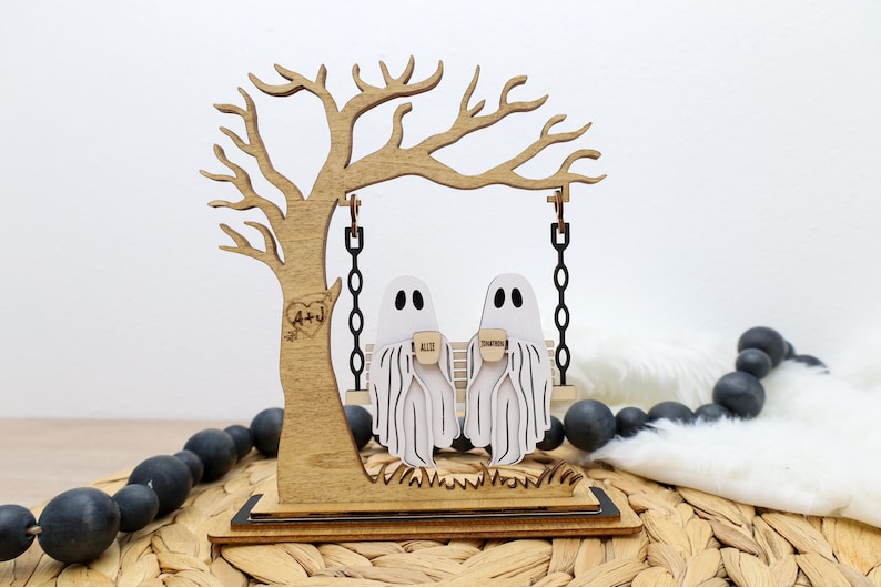 May include: Wooden Halloween decoration featuring a tree with a swing. Two ghost figures are seated on the swing, holding mugs. The tree has a heart with initials carved into it. The base is on a woven surface.