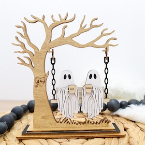 May include: Wooden Halloween decoration featuring a tree with a swing. Two white ghost figures are seated on the swing, holding mugs. The tree has a heart with initials carved into it. The word "TOGETHER" is on the mugs.