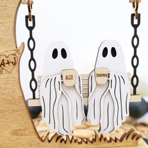 May include: Wooden Halloween decoration featuring two white ghost figures on a swing, each holding a small mug. The mugs are labeled "Allie" and "Jonathon". A wooden tree with a heart and initials is on the left.