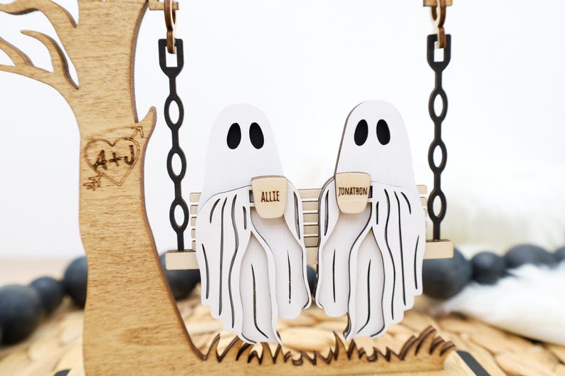 May include: Wooden Halloween decoration featuring two white ghost figures on a swing, each holding a small mug. The mugs are labeled "Allie" and "Jonathon". A wooden tree with a heart and initials is on the left.