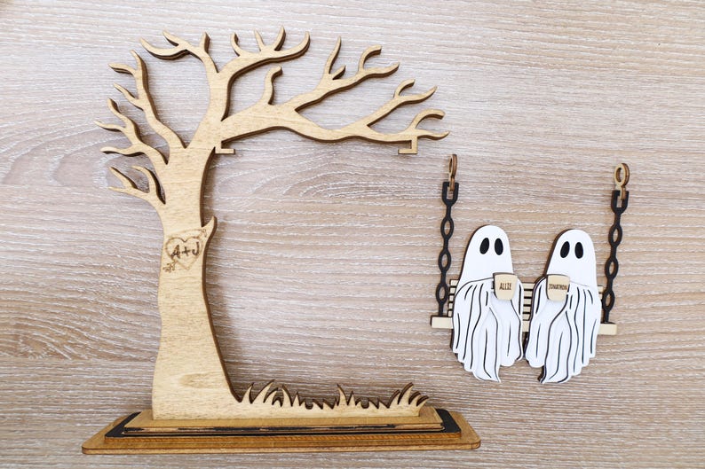 May include: Wooden Halloween-themed decor featuring a tree with initials and a heart, and two ghost figures on a swing. The ghosts hold mugs with the words "All" and "Together". The tree has a base with grass.