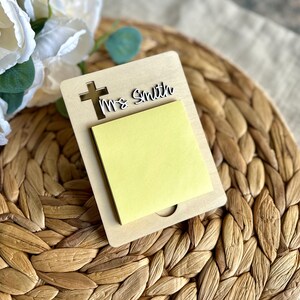 May include: A wooden memo pad holder with a cross cutout and the name "Ms Smith". The holder contains a yellow sticky note pad. The background features a woven placemat and white flowers.