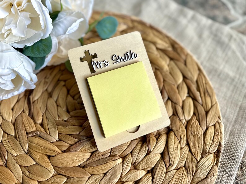 May include: A wooden memo pad holder with a cross cutout and the name "Ms Smith". The holder contains a yellow sticky note pad. The background features a woven placemat and white flowers.