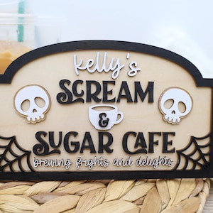 May include: Wooden sign with the text "Kelly's Scream Sugar Cafe" featuring a skull on each side and a coffee cup in the center. The sign has a black border and a light wood background, with the words "brewing frights and delights" at the bottom.
