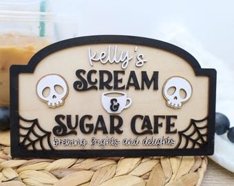 May include: Wooden sign with the text "Kelly's Scream Sugar Cafe" featuring a skull on each side and a coffee cup in the center. The sign has a black border and a light wood background, with the words "brewing frights and delights" at the bottom.