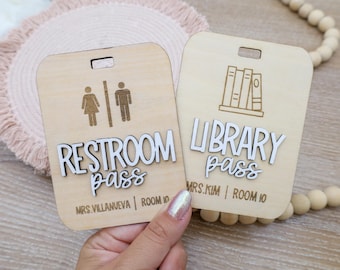May include: Two rectangular light-colored wooden passes with rounded corners and top slots. One reads RESTROOM PASS with a gender icon, the other LIBRARY PASS with a book icon. Both feature white 3D lettering and engraved teacher names and room numbers.